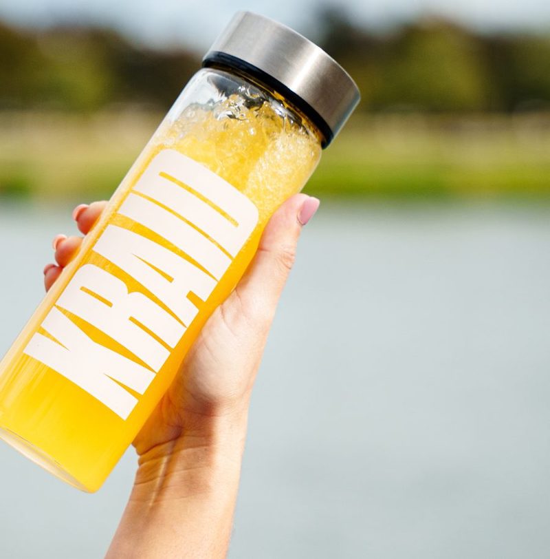 Eine Hand hält eine geschüttelte KRAID-Flasche vor einem See hoch